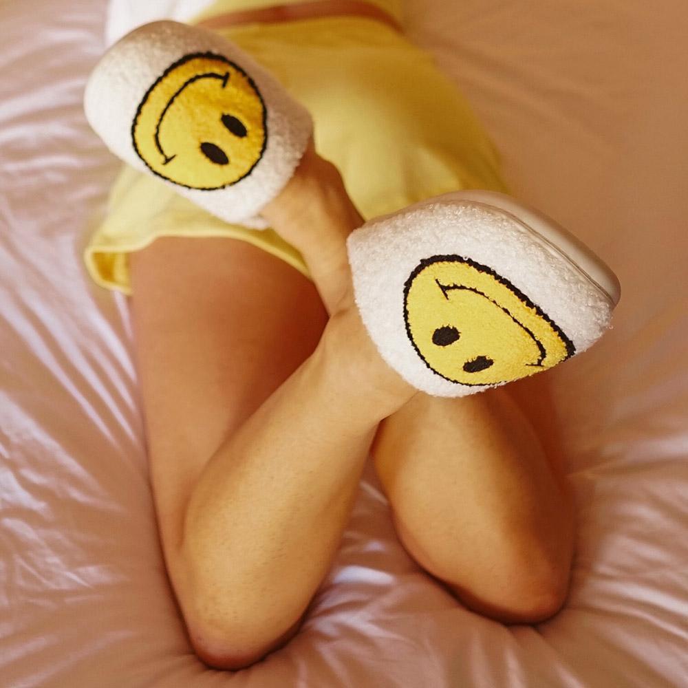 White Fuzzy Happy Face Slippers worn by model laying on bed on tummy